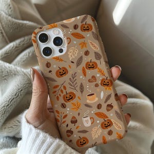 May include: A tan phone case featuring a fall-themed pattern. The design includes orange pumpkins, autumn leaves, coffee cups, and acorns. The case is held in a person's hand, showcasing the detailed print.