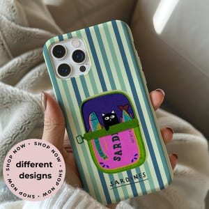 May include: A phone case with a sardine can design. The case has a blue and green striped background. The sardine can is purple with a pink label that reads "Sardines". Inside the can are two fish and a black cat. The text "different designs" is also visible.