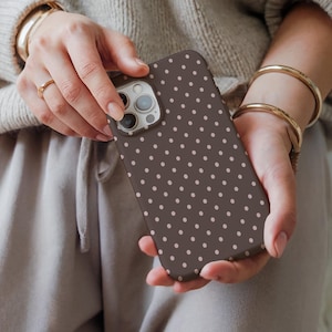 May include: A brown phone case with a repeating pattern of small, light pink polka dots. The phone case is held in two hands, with the camera and buttons visible. The case is designed to fit a smartphone.