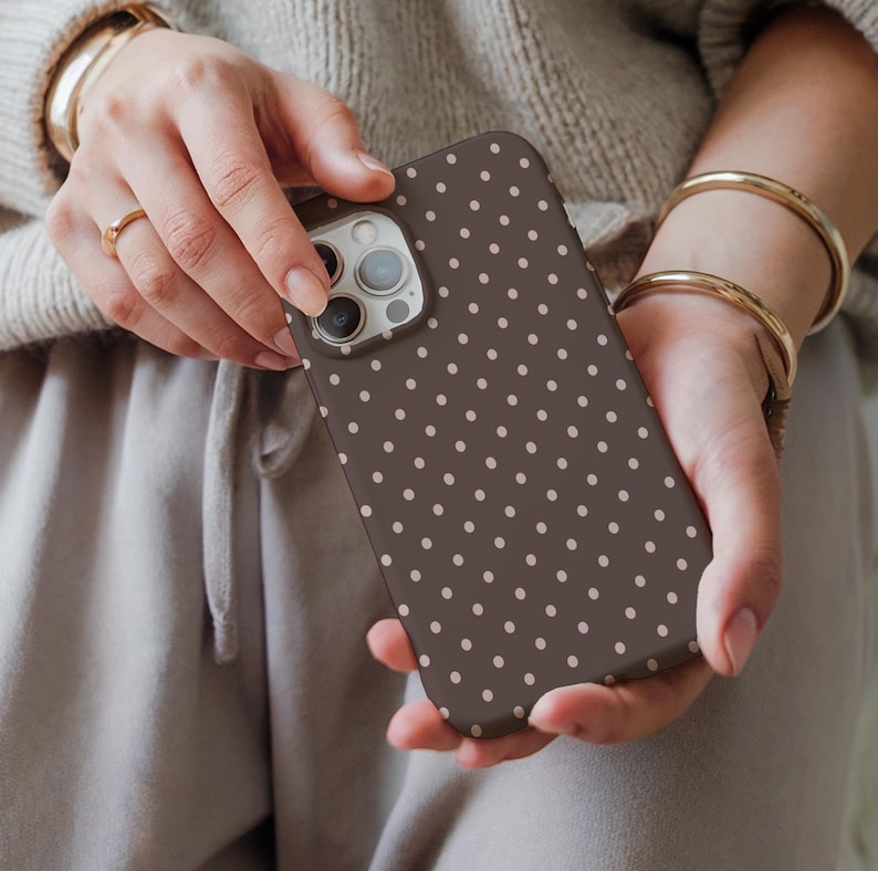 May include: A brown phone case with a repeating pattern of small, light pink polka dots. The phone case is held in two hands, with the camera and buttons visible. The case is designed to fit a smartphone.