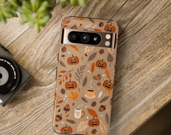 May include: A phone case with a fall-themed pattern featuring pumpkins, leaves, coffee cups, and acorns on a tan background. The case is on a wooden surface, with a camera and greenery in the background.
