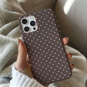 May include: A brown phone case with a repeating pattern of small, light pink polka dots. The case has a camera cutout and is held in a hand with manicured nails. The phone case is designed to protect the phone.