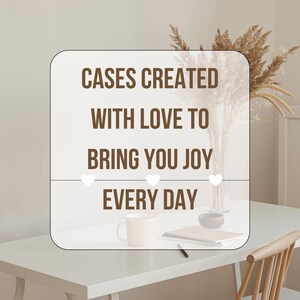 May include: A white rectangular sign with rounded corners displays the text "CASES CREATED WITH LOVE TO BRING YOU JOY EVERY DAY" in brown. Three white heart icons are below the text. A white mug, a small bowl, and a notebook are on a white table.