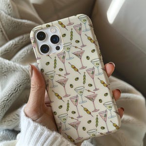 May include: A phone case with a repeating pattern of martini glasses and olives on a light beige background. The martini glasses are in various colors, including pink and green. The olives are on skewers.