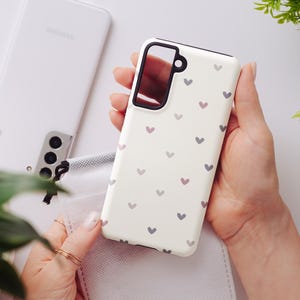 May include: A white phone case with a pattern of small, gray and pink hearts. The case has a black border around the camera cutout. A white Samsung phone and a small white pouch are in the background.
