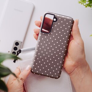 May include: A gray phone case with a white polka dot pattern is held in a person's hands. The case has a black border around the camera and a black accent on the side. A white Samsung phone is in the background.