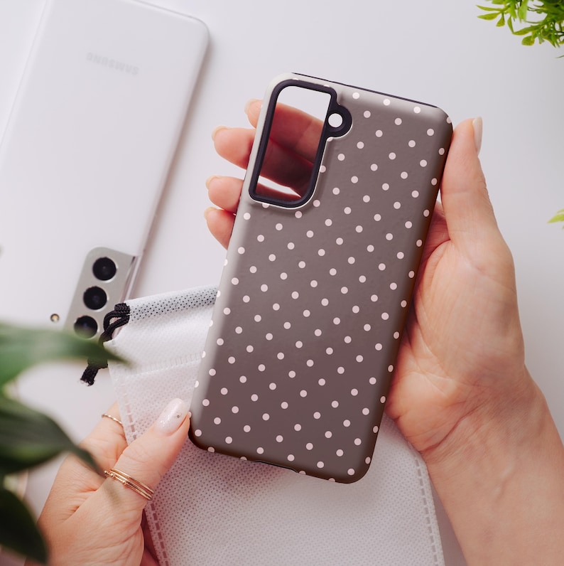 May include: A gray phone case with a white polka dot pattern is held in a person's hands. The case has a black border around the camera and a black accent on the side. A white Samsung phone is in the background.
