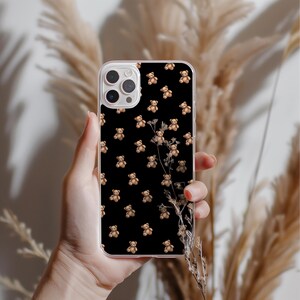 May include: A clear phone case featuring a repeating pattern of small brown teddy bears on a black background. The phone case is held in a hand, with dried plants in the background.
