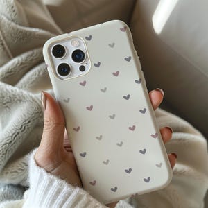 May include: A light-colored phone case with a pattern of small hearts in shades of purple and gray. The case has a camera cutout and button covers. The phone case is held in a person's hand.