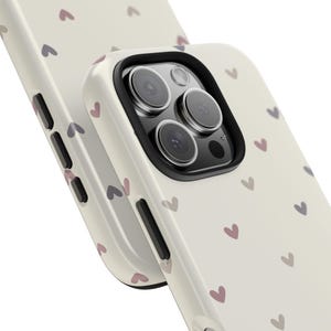 May include: Off-white phone case with a repeating pattern of small, pink and purple hearts. The case has a black camera lens border and a glossy finish. The design is simple and elegant, suitable for everyday use.