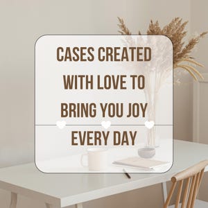 May include: A white rectangular sign with rounded corners displays the text "CASES CREATED WITH LOVE TO BRING YOU JOY EVERY DAY" in brown. Three white hearts are below the text. A mug, bowl, and book are on the table.