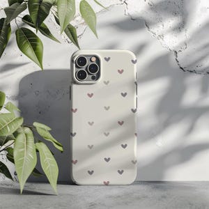 May include: A cream-colored phone case adorned with a pattern of small, heart-shaped designs in shades of purple and pink. The case has a camera module with multiple lenses and a sleek, modern design. The background is a textured, light-colored wall.