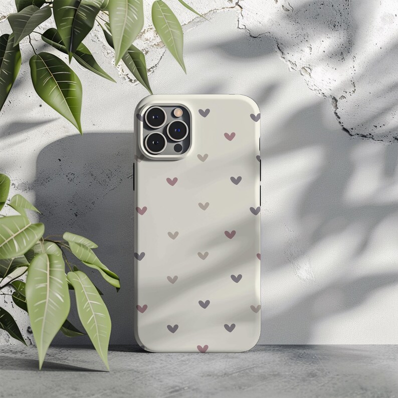 May include: A cream-colored phone case adorned with a pattern of small, heart-shaped designs in shades of purple and pink. The case has a camera module with multiple lenses and a sleek, modern design. The background is a textured, light-colored wall.