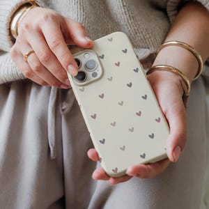 May include: A cream-colored phone case adorned with a pattern of small hearts in shades of purple and gray. The case has a camera cutout and is held in hands.