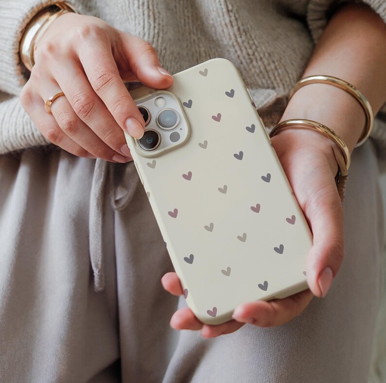May include: A cream-colored phone case adorned with a pattern of small hearts in shades of purple and gray. The case has a camera cutout and is held in hands.