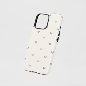 May include: A cream-colored phone case with a pattern of small, evenly spaced hearts in shades of pink and gray. The case has a black border around the camera cutout and the edges. The buttons on the side are black.
