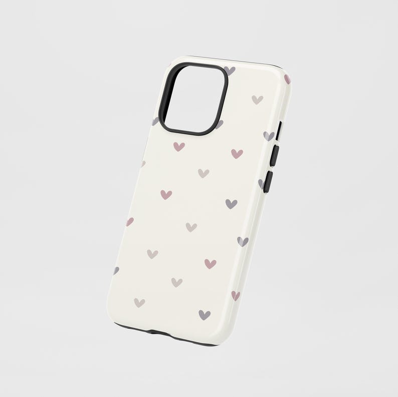 May include: A cream-colored phone case with a pattern of small, evenly spaced hearts in shades of pink and gray. The case has a black border around the camera cutout and the edges. The buttons on the side are black.