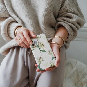 Könnte beinhalten: Eine weiße Handyhülle mit einem Blumendesign mit weißen Lilien und grünen Blättern.