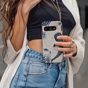 May include: A smartphone case with a marine life design. The case features a whale shark, coral, and other sea creatures in shades of blue and gray. The case is held by a person wearing a black top and blue jeans.