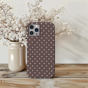 May include: A brown phone case with a repeating pattern of small, light pink polka dots. The case is shown on a wooden surface, with a vase of white flowers in the background.