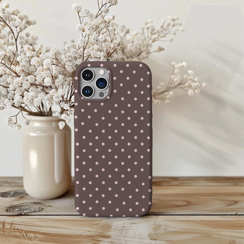 May include: A brown phone case with a repeating pattern of small, light pink polka dots. The case is shown on a wooden surface, with a vase of white flowers in the background.