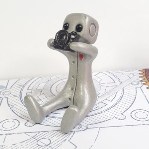 May include: A small, silver, robot figurine holding a black camera. The robot has a red heart on its chest and black eyes. The robot is sitting on a white surface with a blue and white pattern.