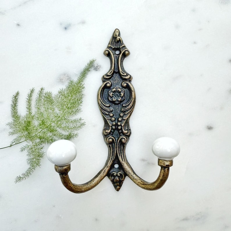 May include: A double hook wall hanger with an ornate, antique gold finish and two white ceramic knobs.