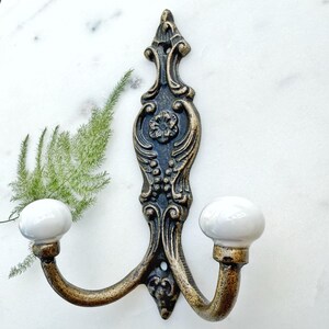 May include: A double hook wall hanger with a decorative ornate design in black and gold with white ceramic knobs.