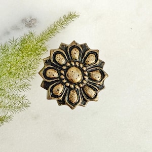 May include: A bronze-colored, flower-shaped decorative knob. The knob has a detailed design with a central circle surrounded by petal-like shapes. The knob is set against a white marble surface, with a sprig of green foliage in the background.