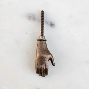 May include: A bronze-colored hand-shaped cabinet knob with a threaded rod for installation. The hand is detailed with visible knuckles and a slightly curved palm. The knob is designed to be mounted on a cabinet or drawer.