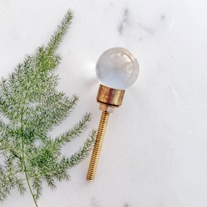 May include: A clear glass knob with a brass base and screw. The knob is round and has a smooth surface. The screw is threaded and has a gold finish.