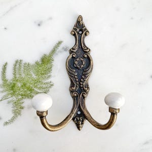 May include: A double hook wall mount with an ornate, gold-toned metal design and two white ceramic knobs.