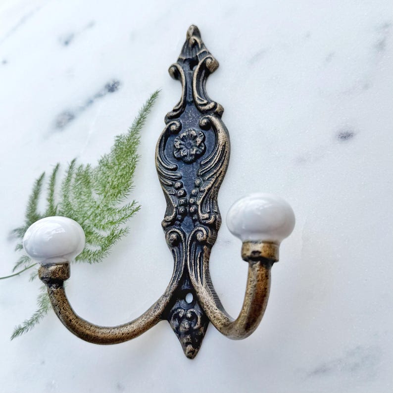 May include: Ornate double hook with a decorative scroll design in black and gold with two white ceramic knobs.