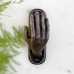 May include: Bronze-toned hand-shaped door knocker with a detailed design. The knocker is mounted on an oval backplate with a beaded border. The hand is in a welcoming gesture, with visible fingernails.