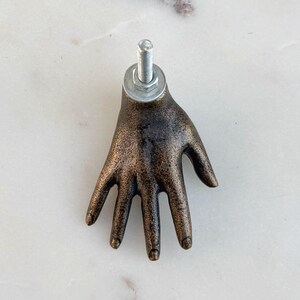 May include: A bronze-colored hand-shaped cabinet pull with a bolt and washer attached. The hand is detailed with visible knuckles and a textured surface, suggesting an antique or vintage aesthetic. The hardware is metallic and contrasts with the bronze finish.