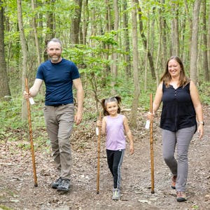 May include: A family of three walks through a forest, each holding a wooden walking stick. The father wears a blue shirt and khaki pants, the mother wears a black tank top and gray pants, and the child wears a purple shirt and black pants.