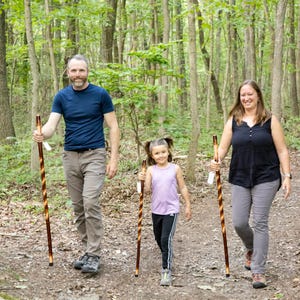 May include: A family of three, a man, a woman, and a young girl, are hiking in a forest. They are all holding walking sticks. The man is wearing a blue shirt and khaki pants. The woman is wearing a black tank top and gray pants. The girl is wearing a purple shirt and black pants.