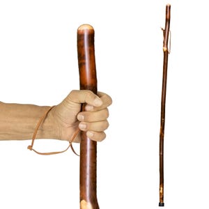 May include: A wooden walking stick with a brown finish and a leather wrist strap. The stick is held in a hand, showcasing its polished surface and natural wood grain. A second stick is shown to the right, with a leather strap near the top.