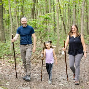 May include: A family of three hikes through a wooded area. The father wears a blue shirt and khaki pants, the mother wears a black tank top and gray pants, and the daughter wears a purple shirt and black pants. All three are holding walking sticks.