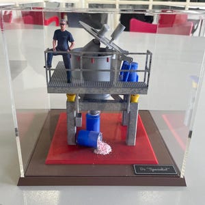 Peut inclure: Maquette industrielle miniature dans un présentoir transparent. Le modèle présente une machine à mélanger argentée avec une figurine d'une personne sur une plateforme. Un baril bleu est renversé, déversant de la poudre rose. La base est rouge avec une bordure marron.