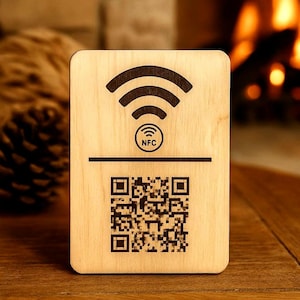 Pode incluir: Uma placa retangular de madeira clara com um símbolo de Wi-Fi gravado em castanho escuro no topo, um pequeno círculo com o texto NFC no centro e um grande código QR quadrado impresso na metade inferior da superfície de madeira.