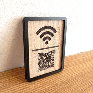 Può includere: Un cartello rettangolare in legno con cornice nera e centro in legno chiaro. Il cartello presenta un simbolo Wi-Fi inciso in scuro nella parte superiore e un codice QR sottostante, separati da una linea orizzontale. Il design è centrato sul legno.