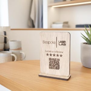 May include: A wooden sign with the text "Bespoke Laser Lab" and "Leave us a Review" with a QR code and star rating. The sign is light brown and stands on a wooden surface.