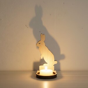 May include: Wooden bunny candle holder with a lit white tea light. The bunny silhouette is light brown, with a dark brown base. The shadow of the bunny is cast on the wall behind it. A decorative item for Easter or spring.