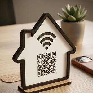 Könnte beinhalten: Ein holzgeschild in Hausform mit schwarzer Umrandung und Sockel. Das Schild hat eine weiße Mitte mit einem schwarzen Wi-Fi-Symbol und einem QR-Code. Eine kleine Sukkulentenpflanze im Topf und ein Telefon befinden sich im Hintergrund.