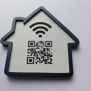Może przedstawiać: Drewniany podkładka w kształcie domu z czarną obwódką. Podkładka ma białe wnętrze z kodem QR i symbolem Wi-Fi. Projekt jest prosty i nowoczesny, odpowiedni do użytku domowego lub biurowego.