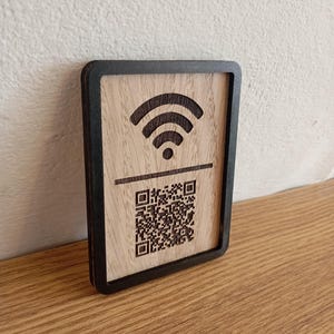 May include: Wooden WiFi sign with a black border. The sign features a carved WiFi symbol and a QR code. The wood has a natural grain, and the sign is rectangular. The sign is designed to provide WiFi access.