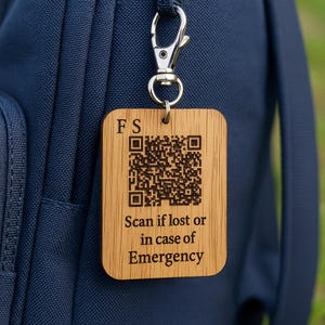 Könnte beinhalten: Ein Holzanhänger mit einem QR-Code und dem Text "Scan if lost or in case of Emergency" ist an einem Rucksack befestigt. Der Anhänger hat die Initialen "FS" oben. Der Anhänger ist rechteckig mit abgerundeten Ecken und wird mit einem silbernen Verschluss befestigt.