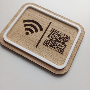 Può includere: Un cartello rettangolare in legno con angoli arrotondati e un bordo bianco in rilievo. La superficie è incisa con un'icona Wi-Fi nera sulla sinistra e un codice QR quadrato sulla destra, separati da una sottile linea verticale.
