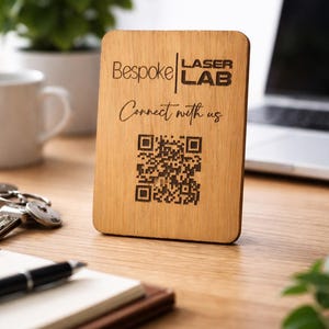 Könnte beinhalten: Ein rechteckiges Holzschild mit dem Text "Bespoke Laser Lab" und "Connect with us" in einer handschriftlichen Schriftart. Unter dem Text befindet sich ein QR-Code. Das Schild steht auf einem Holztisch mit einem Laptop und einer Pflanze im Hintergrund.
